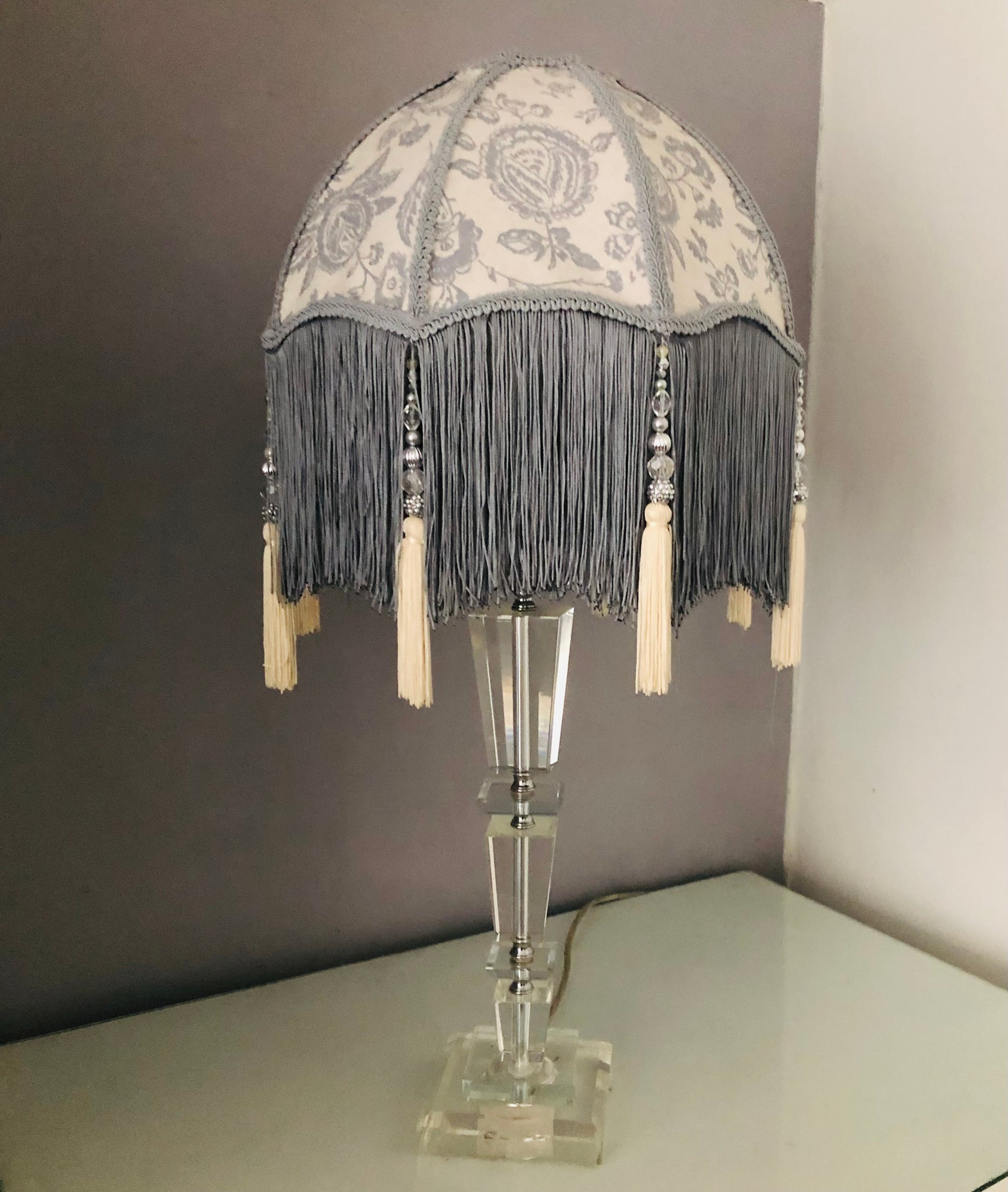 Full lampshade view – A grey Victorian parachute-style lampshade in blue toile fabric, with a soft scalloped shape and a long beaded fringe on.glass lamp