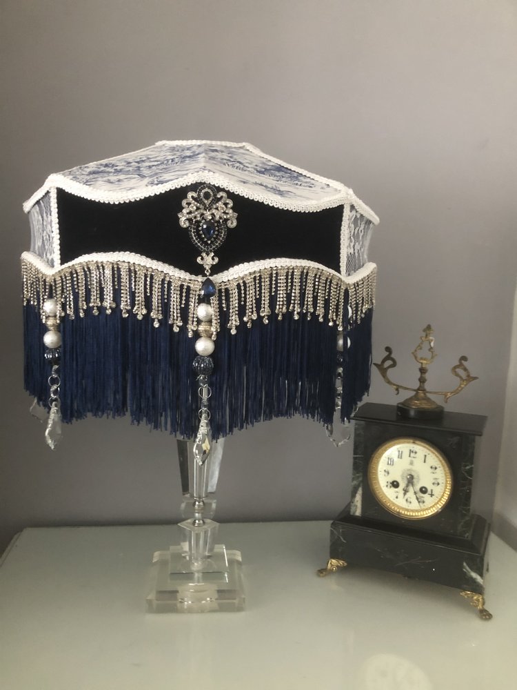 victorian_fringed_lampshade, in blue velvet toile and lace, with diamantes and blue fringe on a  glass lamp  next  to an antique clock. two