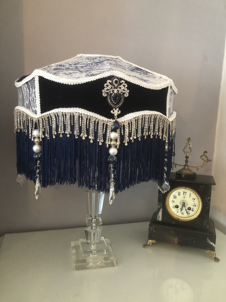 victorian_fringed_lampshade, in blue velvet toile and lace, with diamantes and blue fringe on a  glass lamp  next  to an antique clock. three